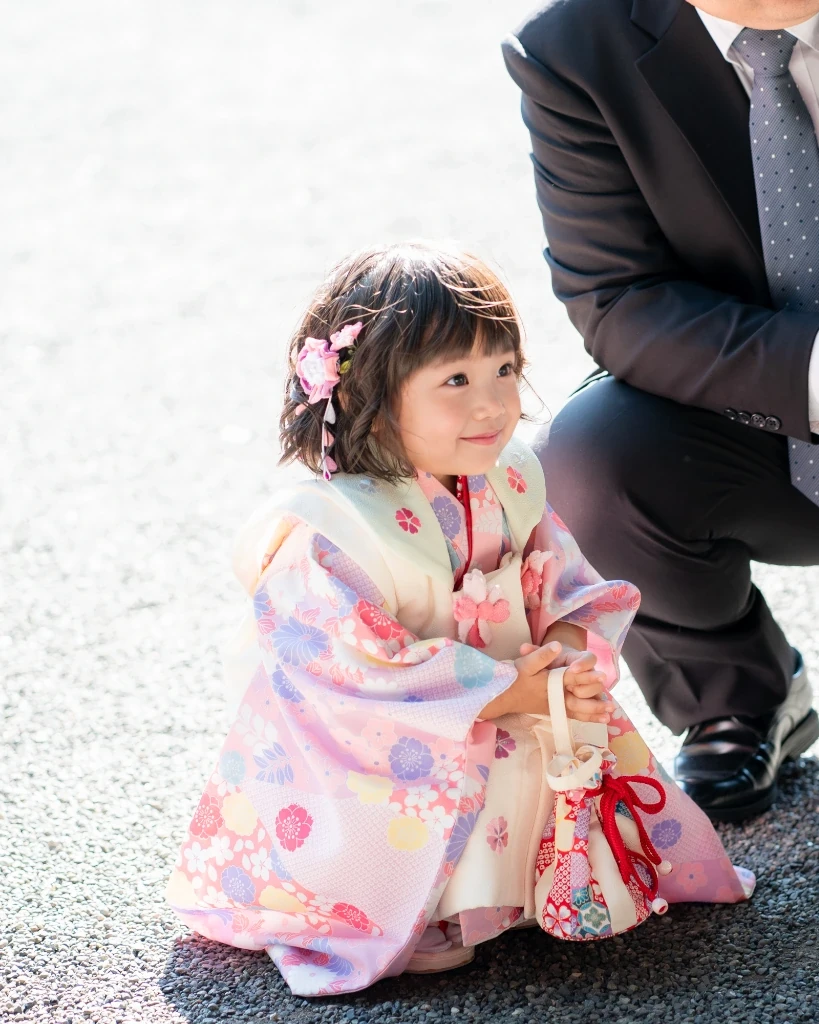 おろしたくせ毛のボブに、ピンクの花と下がり飾りをつけた3歳の女の子がしゃがんで笑う