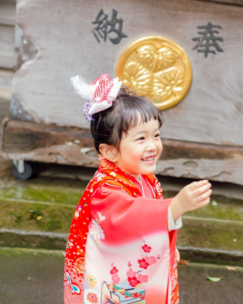 ボブで前髪ありの3歳の女の子が笑顔で手を伸ばす。髪をトップにまとめ、羽根つきの髪飾りをつけている