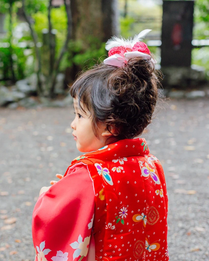 ボブでくせ毛の3歳の女の子を後ろ斜めから。髪をトップにまとめ、ピンクと白の羽根飾りをつけている
