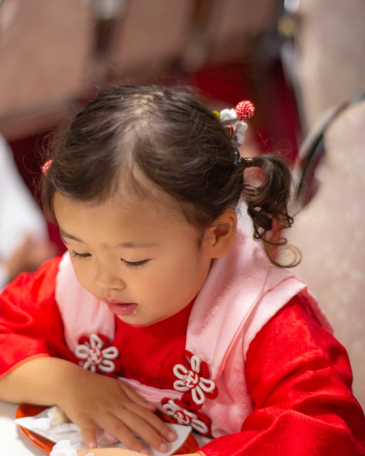 七五三3歳の女の子。短く少ない髪を耳の上で小さな二つ結びにし、赤と白の飾りをつけた被布姿