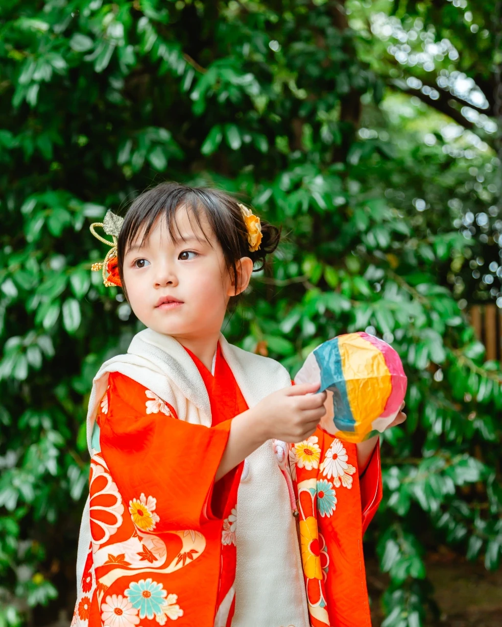 両サイドお団子にした3歳の女の子の横顔。金色の葉と橙色の花の髪飾りをつけ、紙風船を持つ