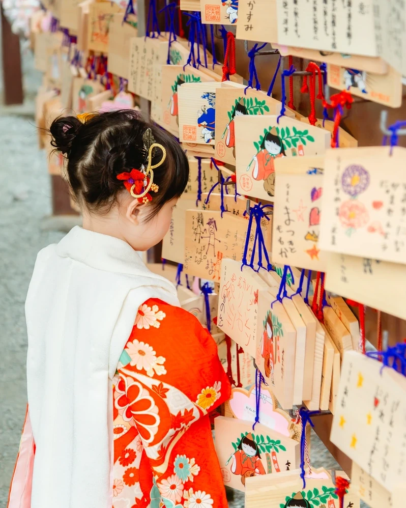 小さなお団子に赤と金の髪飾りをつけた3歳の女の子が、神社で絵馬に手を伸ばす後ろ姿