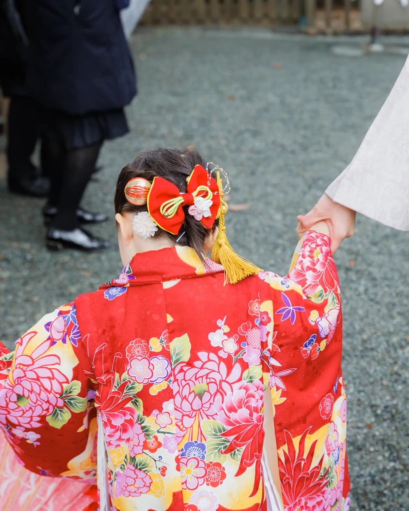 髪をまとめて赤い大きなリボンと金の玉飾りをつけた3歳の女の子が、家族と手をつないで歩く後ろ姿