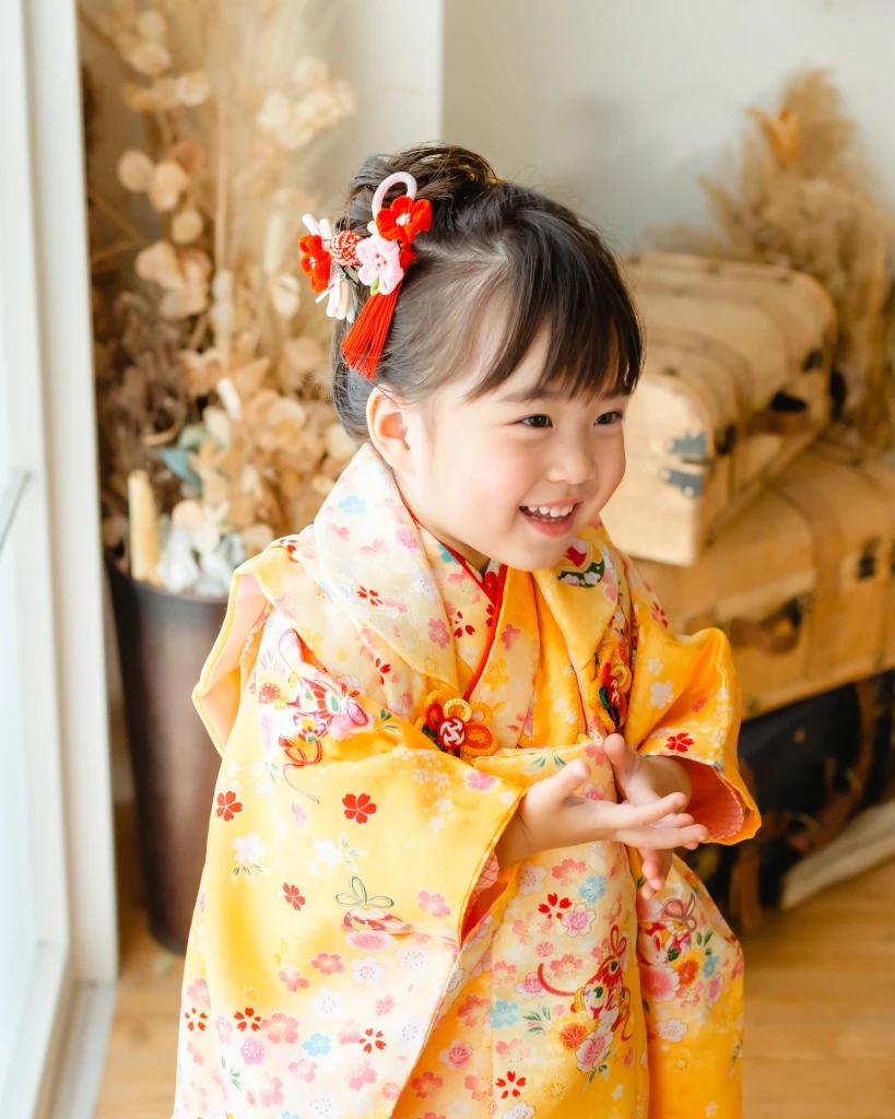 前髪ありの3歳の女の子。髪をサイドにまとめ、赤い花と房の髪飾りをつけた黄色の着物姿で笑う