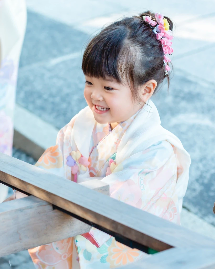 サイドに髪をまとめ、ピンクの下がり飾りをつけた3歳の女の子の横顔。淡い色の被布姿