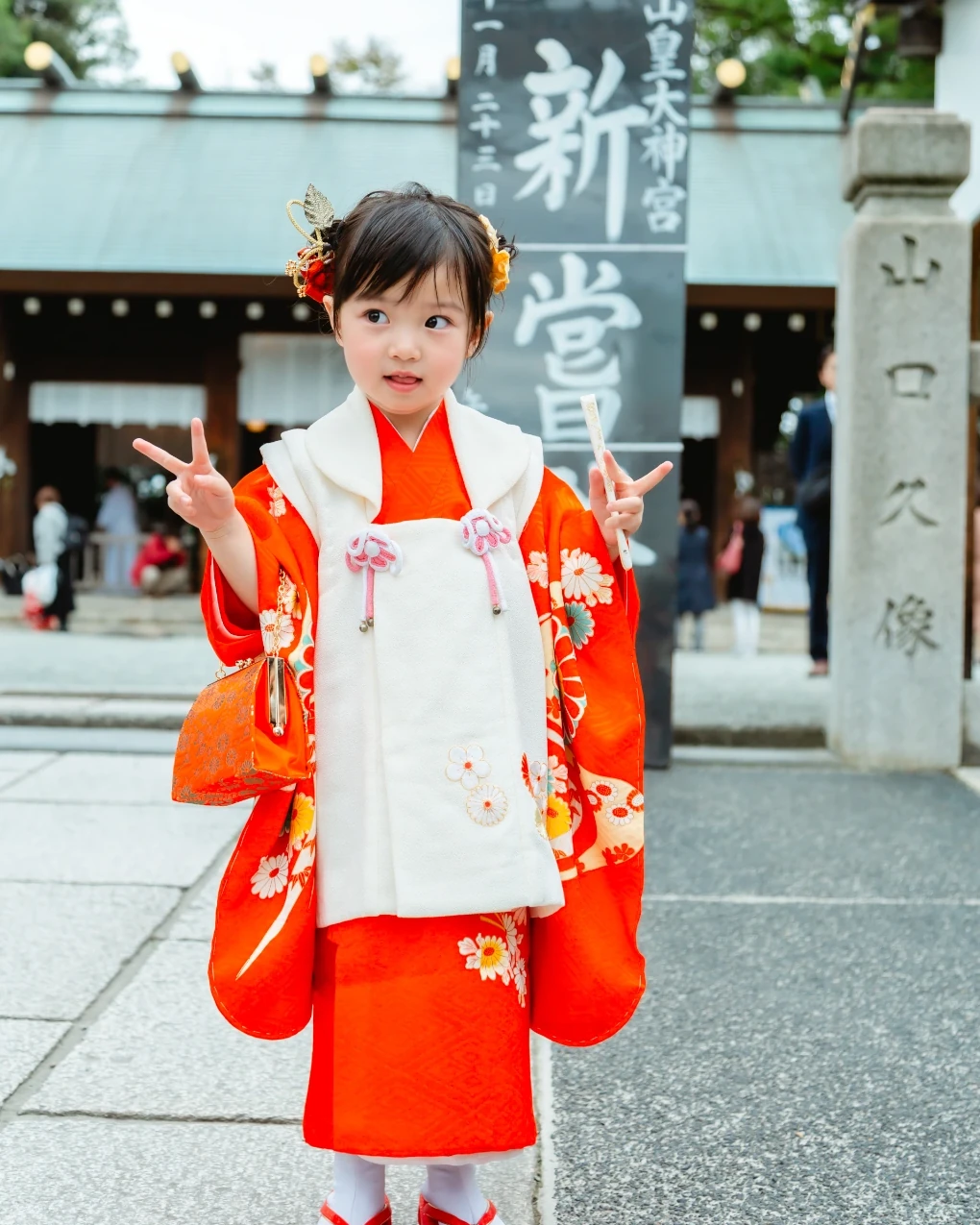 金色の葉と橙色の花の髪飾りをつけた3歳の女の子。赤い着物に白い被布姿で神社の境内に立つ