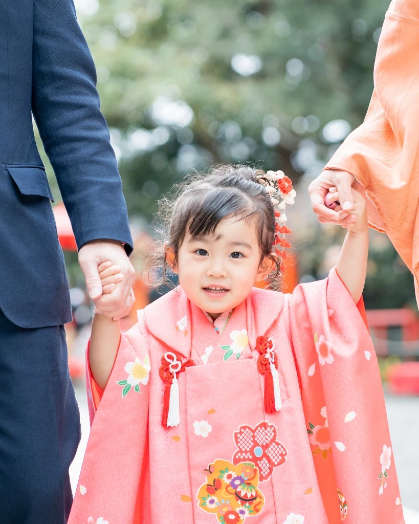 髪をサイドにまとめて花飾りをつけた3歳の女の子が、両親と手をつないで歩く