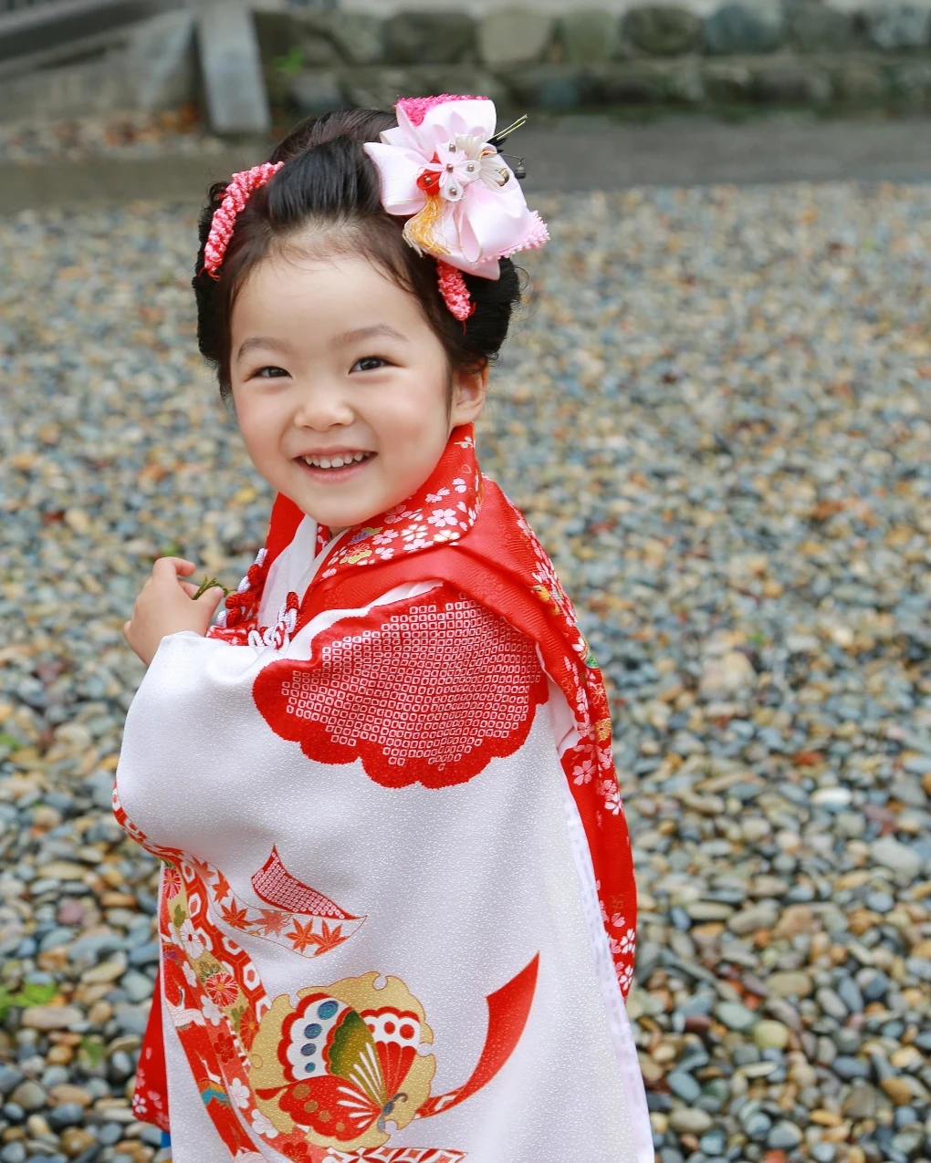 前髪なしでおでこを出した3歳の女の子。髪をサイドにまとめ、大きなピンクの花飾りをつけて笑う