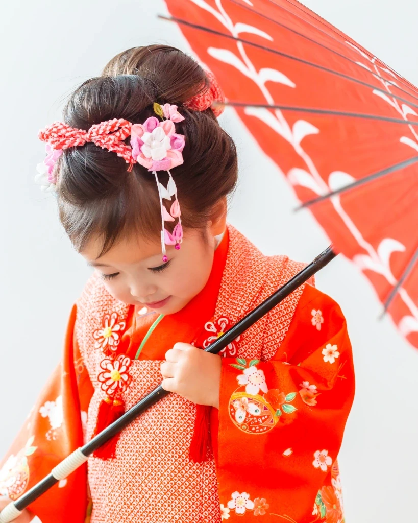 前髪なしの3歳の女の子。髪をトップにまとめ、ピンクの花と下がり飾りをつけた被布姿で和傘を持つ