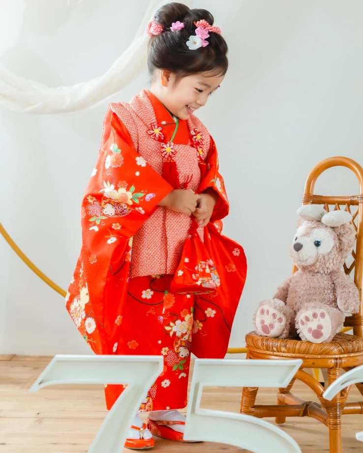 トップにお団子を作り、ピンクの花飾りをつけた3歳の女の子の全身。赤い被布姿でスタジオに立つ