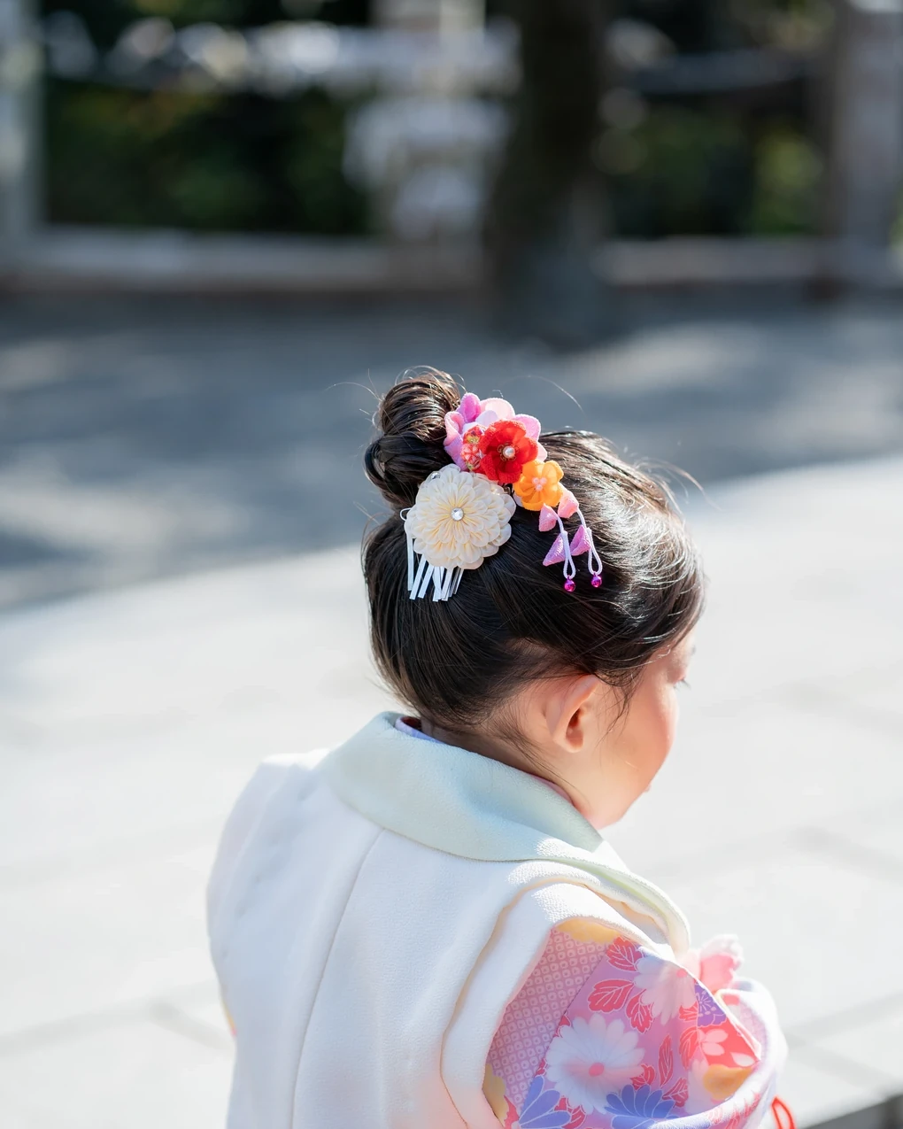 トップにお団子を作った3歳の女の子を後ろ斜めから。白や赤の花と下がり飾りをつけた被布姿