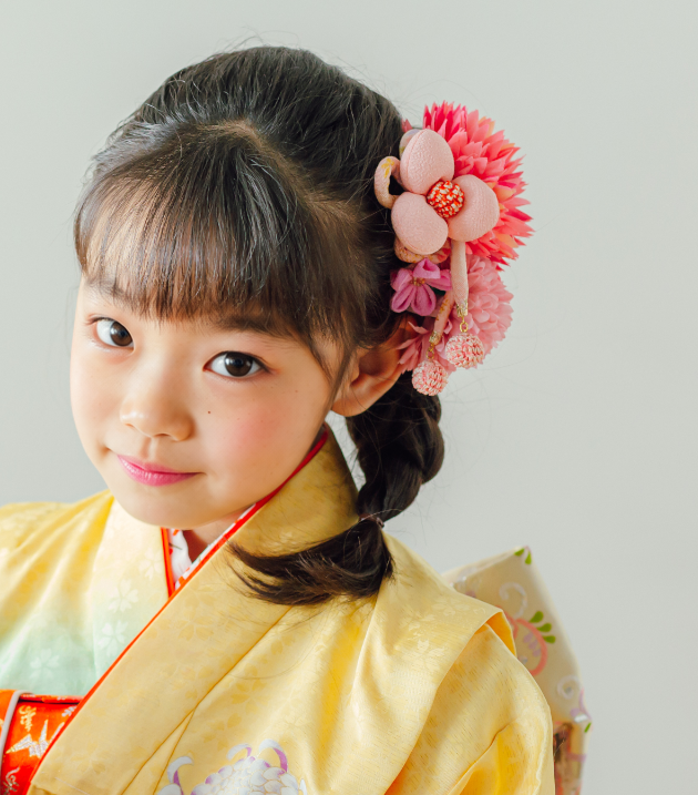 七五三で7歳の女の子の髪型のアップ。ぱっつん前髪のまま、サイドの三つ編みにピンクの大輪の花