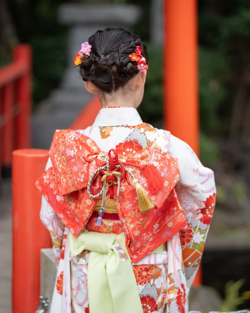 赤い欄干の前で、七五三7歳の後ろ姿。編み込みを低めにまとめたアップに小さな花飾り