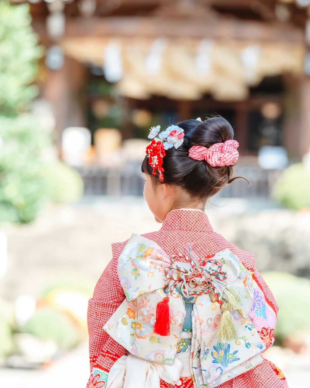 神社の社殿を背にした七五三7歳の後ろ姿。低めのアップにピンクの鹿の子と赤・水色の花飾り