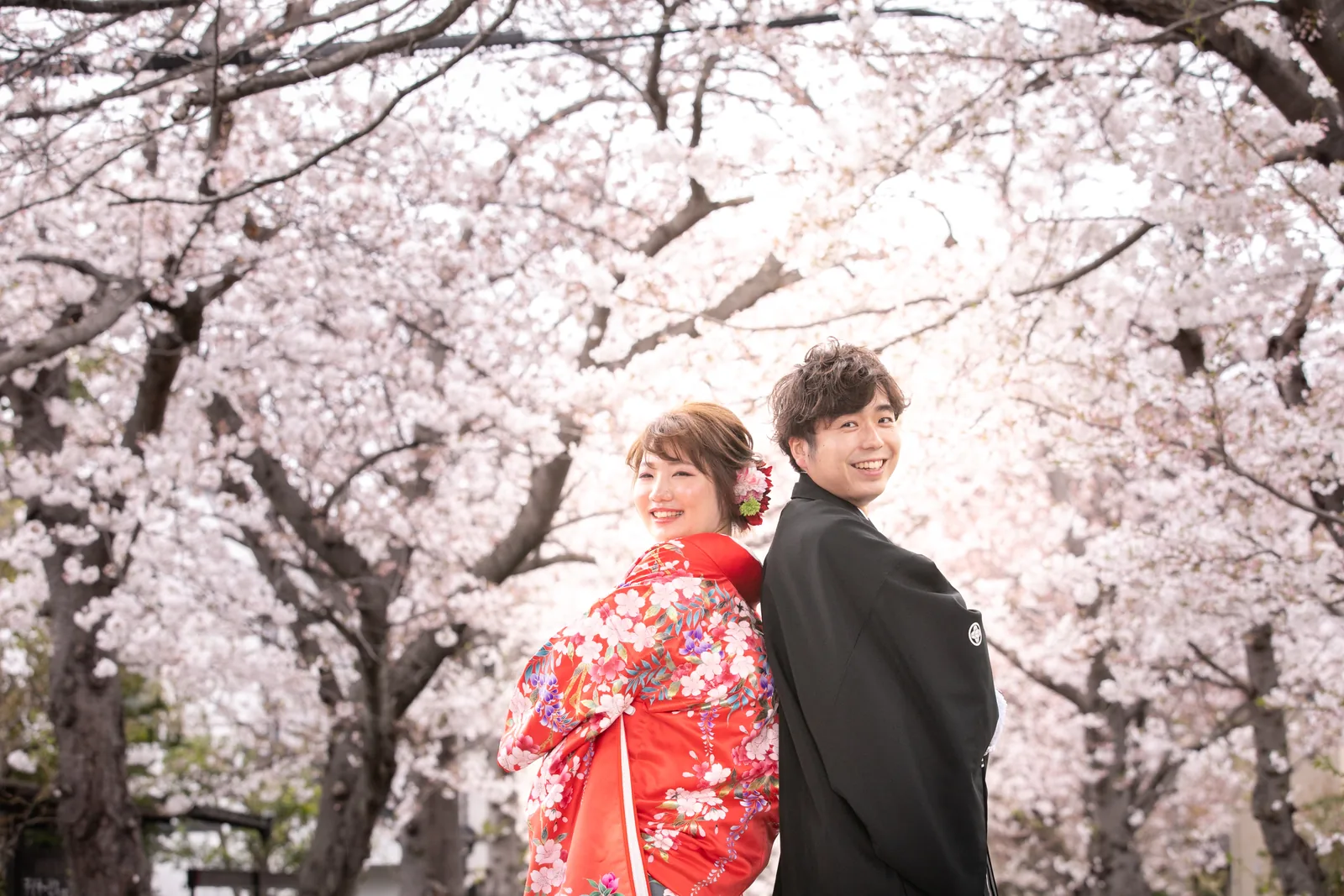 称名寺の桜並木の参道で、色打掛の新婦と紋付袴の新郎が背中合わせに立っている