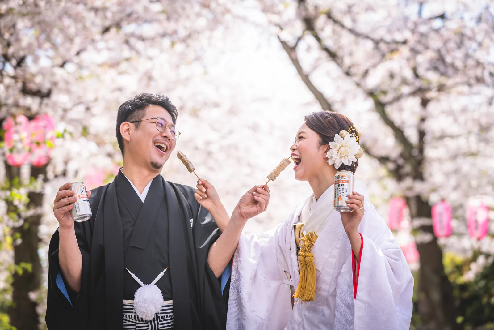 称名寺の桜並木の下で、白無垢の新婦と紋付袴の新郎が顔を見合わせて笑っている