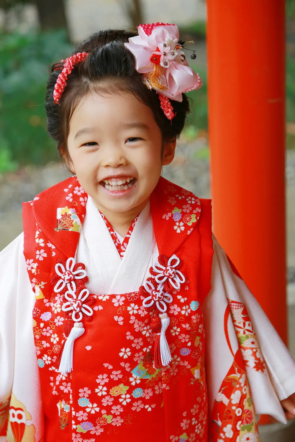 朱塗りの柱を背に歯を見せて笑う3歳の女の子、赤地に桜を散らした被布と日本髪風の結い上げの七五三（相州春日神社・横浜）