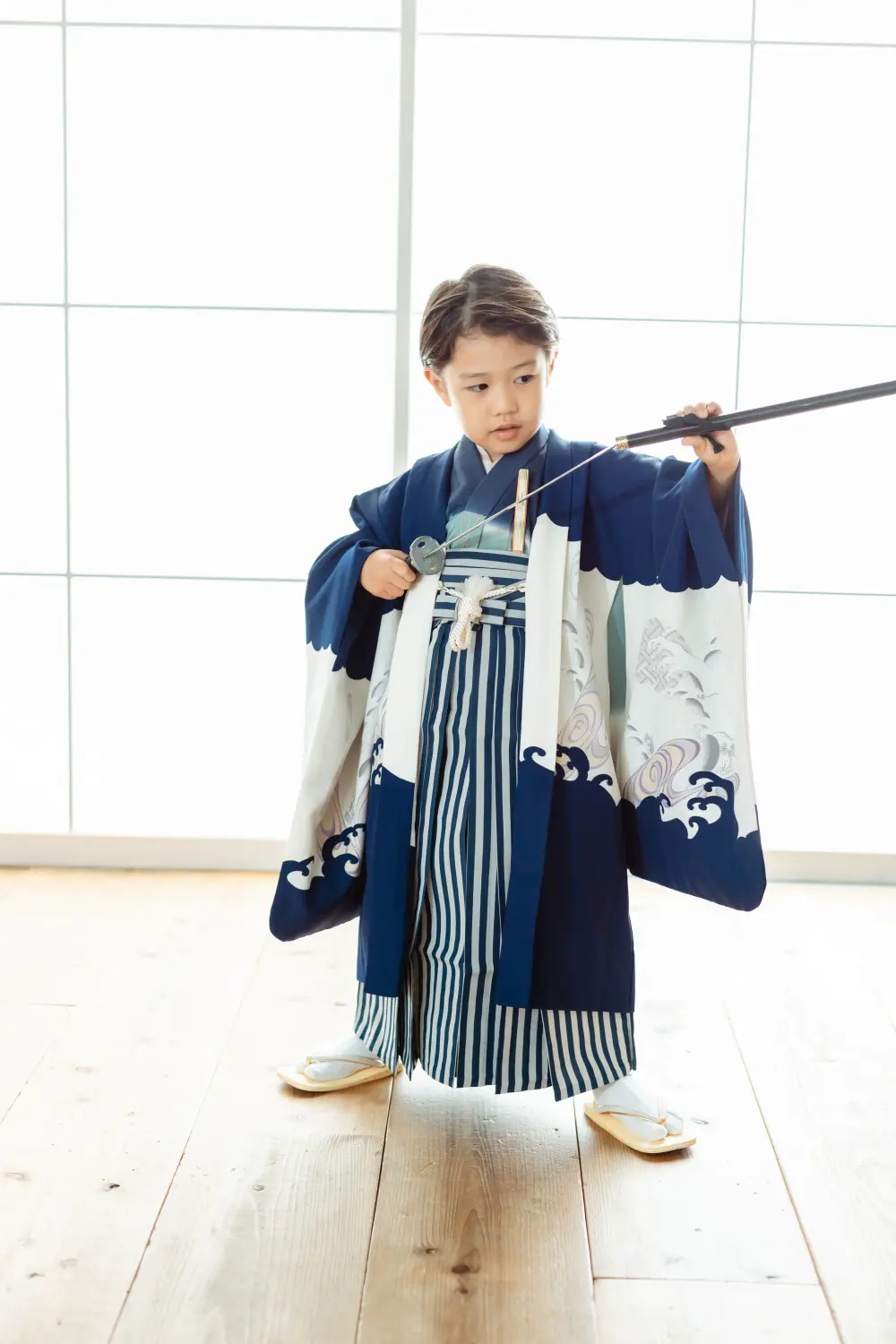 白い格子窓を背に小道具を手にした羽織袴姿の5歳の男の子、七五三スタジオ撮影