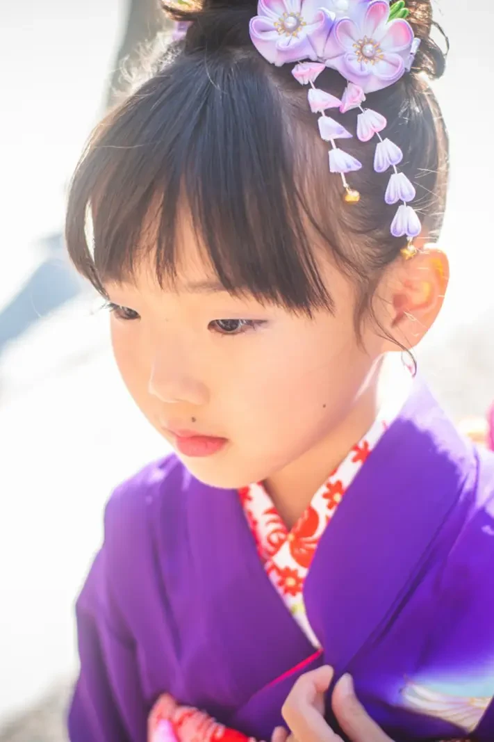 逆光に包まれ伏し目がちにたたずむ紫の四つ身姿の7歳の女の子、七五三（武田神社・甲府）