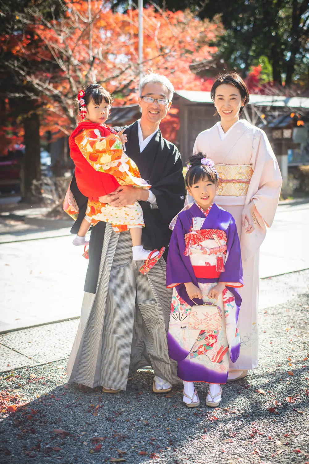 真紅の紅葉を背に父に抱かれる3歳の被布姿の妹と、隣に立つ7歳の四つ身の姉の七五三家族写真（武田神社）