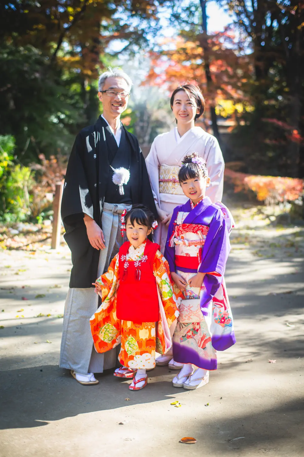黒紋付羽織袴の父と淡いピンクの訪問着の母、7歳と3歳の姉妹が紅葉の参道に並ぶ七五三の家族写真（武田神社）