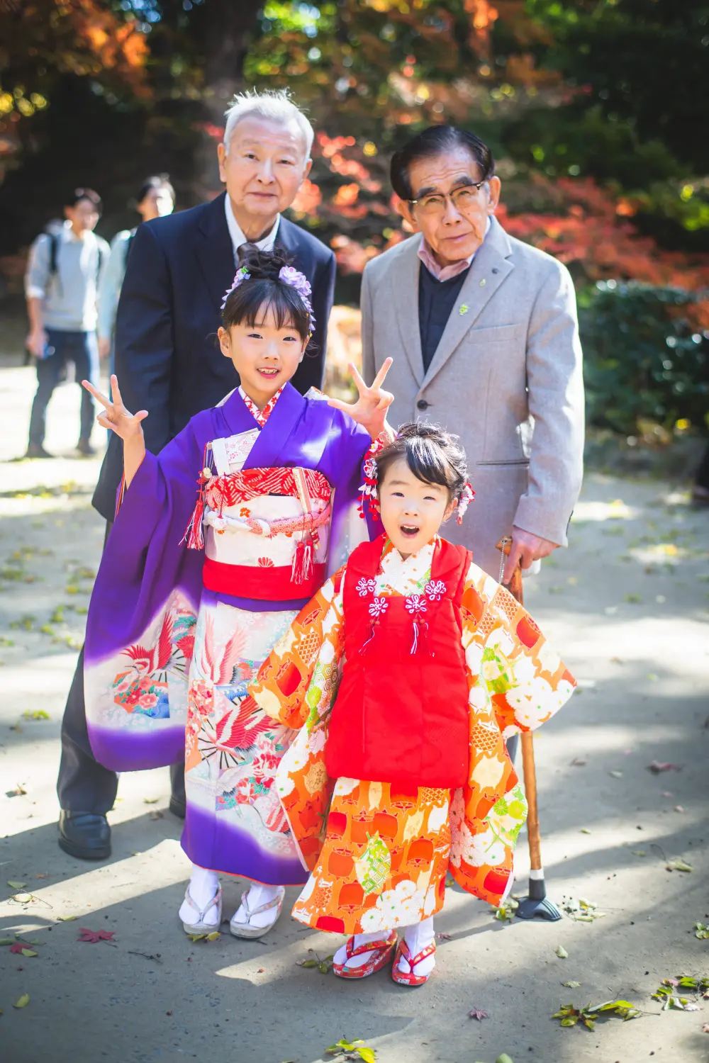 ピースサインをする7歳の四つ身と3歳の被布の姉妹、後ろで見守るご家族、七五三（武田神社）