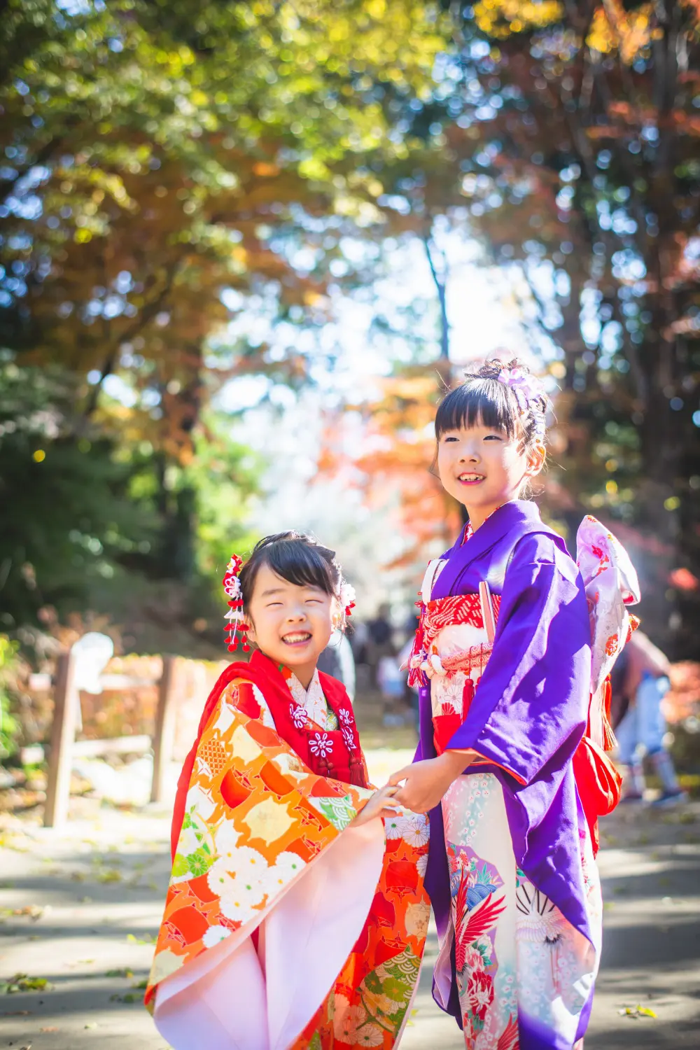木漏れ日の参道で手をつないで笑う7歳の四つ身と3歳の被布の姉妹、七五三（武田神社・山梨）