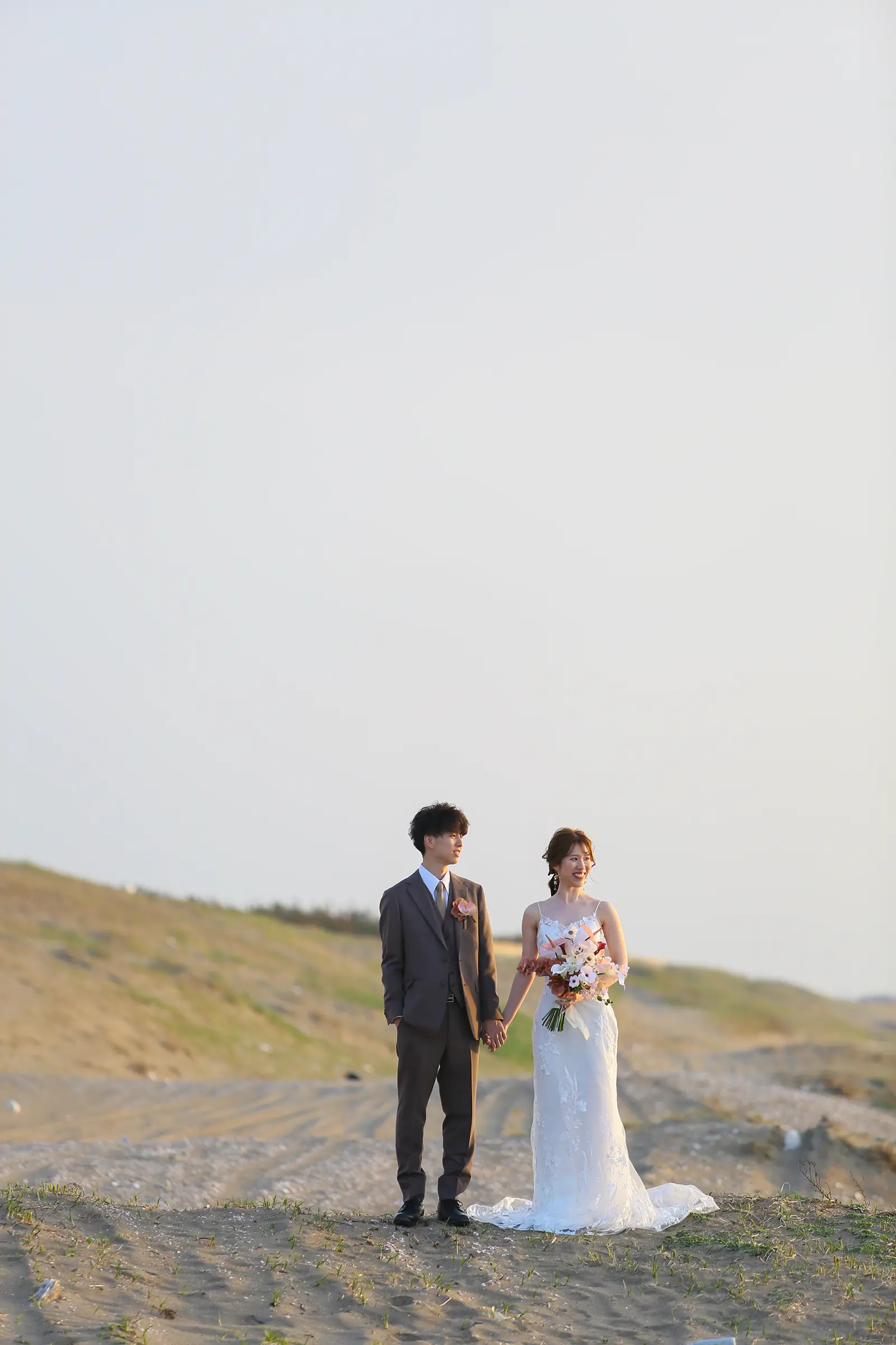 内灘マリーナ周辺の砂丘と海辺で洋装の新郎新婦が手をつないで立つフォトウェディング写真