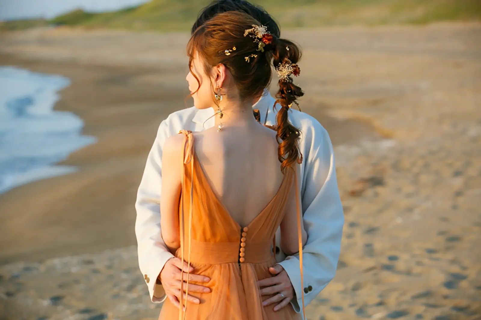 内灘マリーナ周辺の海辺でオレンジのカラードレスを着た新婦と白いタキシードの新郎が手をつないで歩く写真