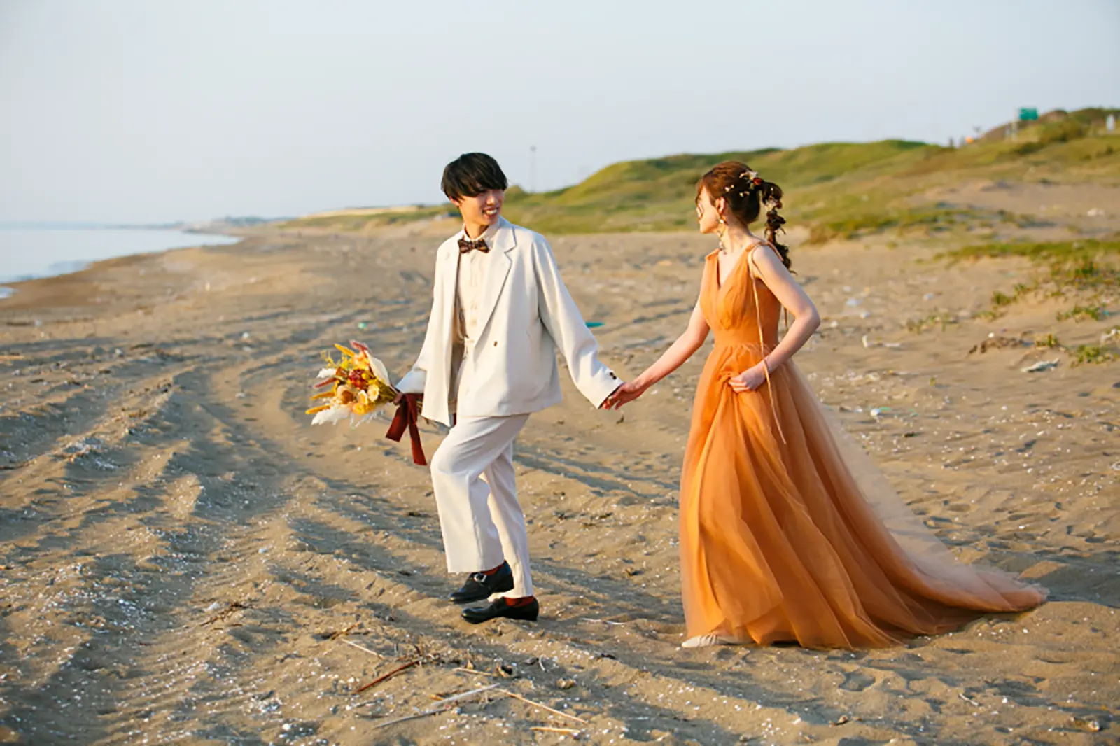 内灘マリーナ周辺の海辺でオレンジのカラードレスを着た新婦と白いタキシードの新郎が手をつないで歩く写真