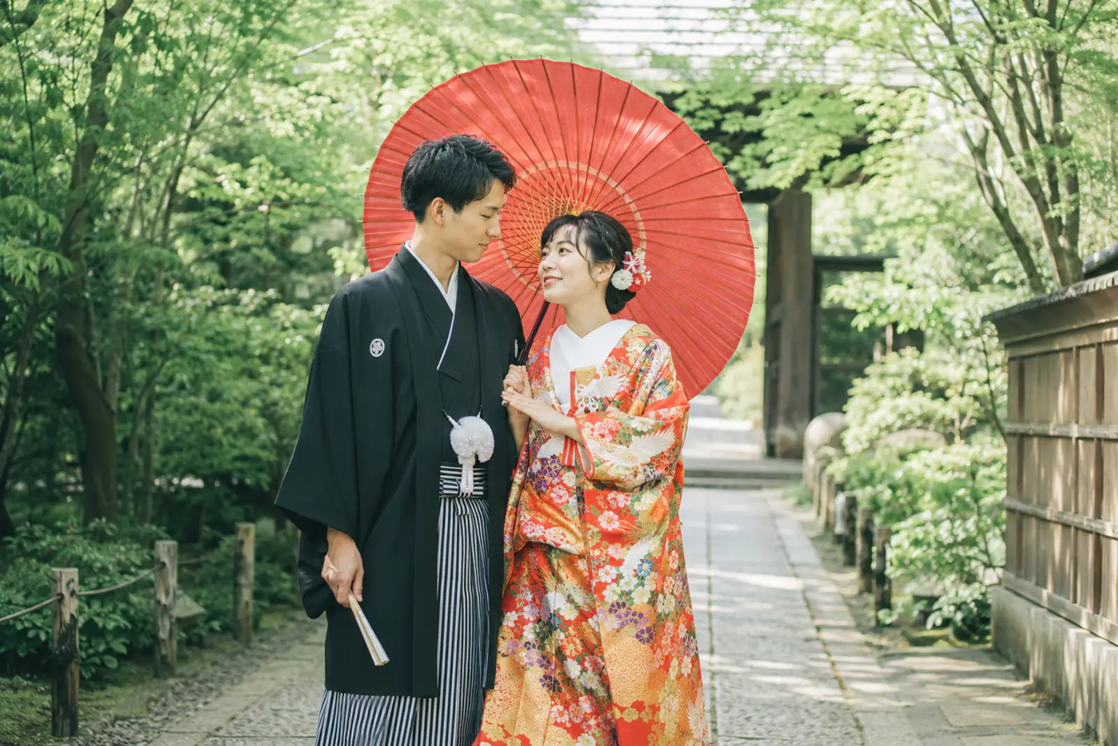 色打掛の新婦と紋付袴の新郎が赤い和傘を使って見つめ合う和装前撮り写真