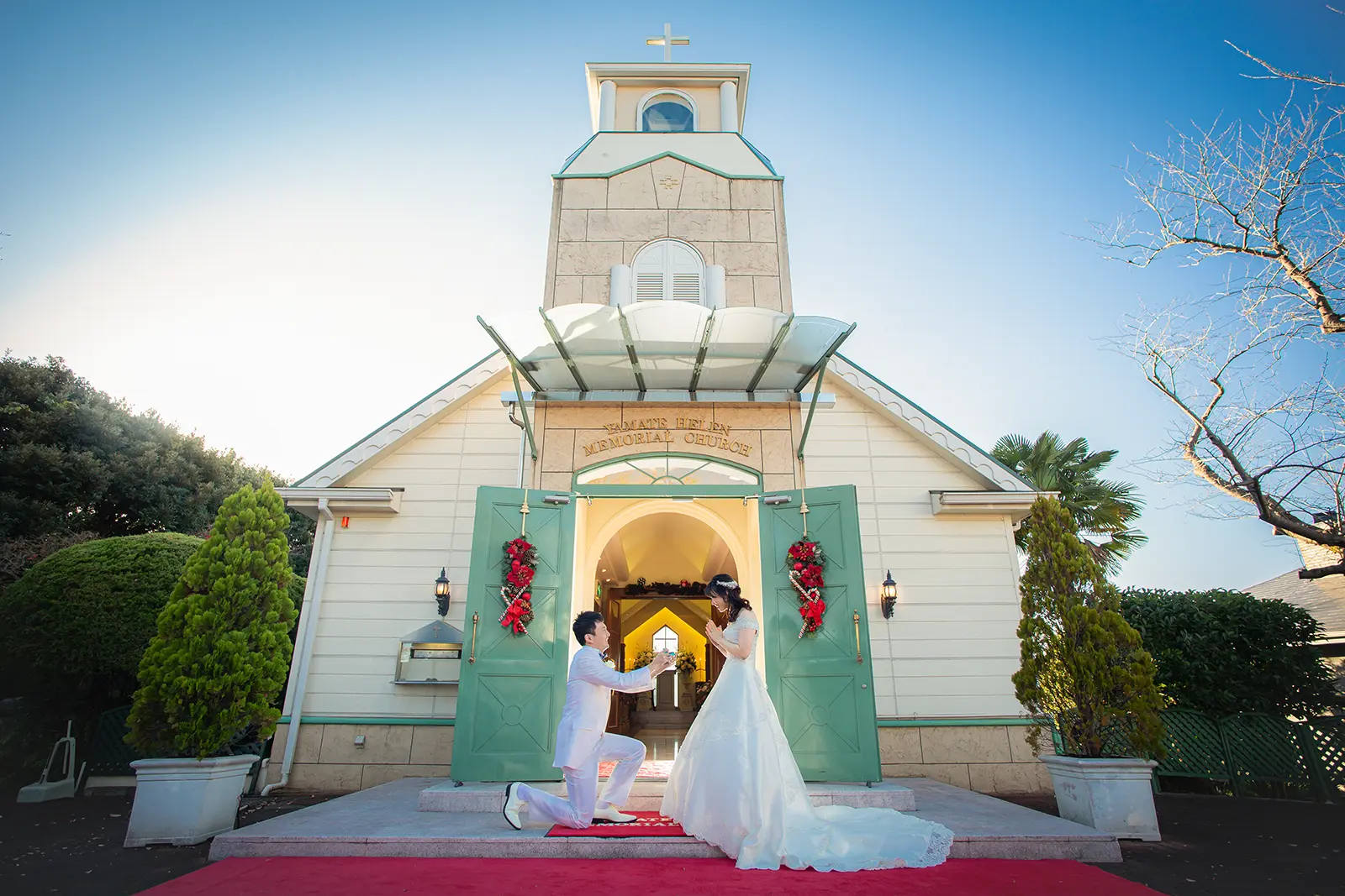 横浜の山手ヘレン記念教会の入り口で、タキシード姿の男性がドレス姿の女性にプロポーズしている瞬間。フォトジェリッシュによるドラマチックなウェディングフォト。