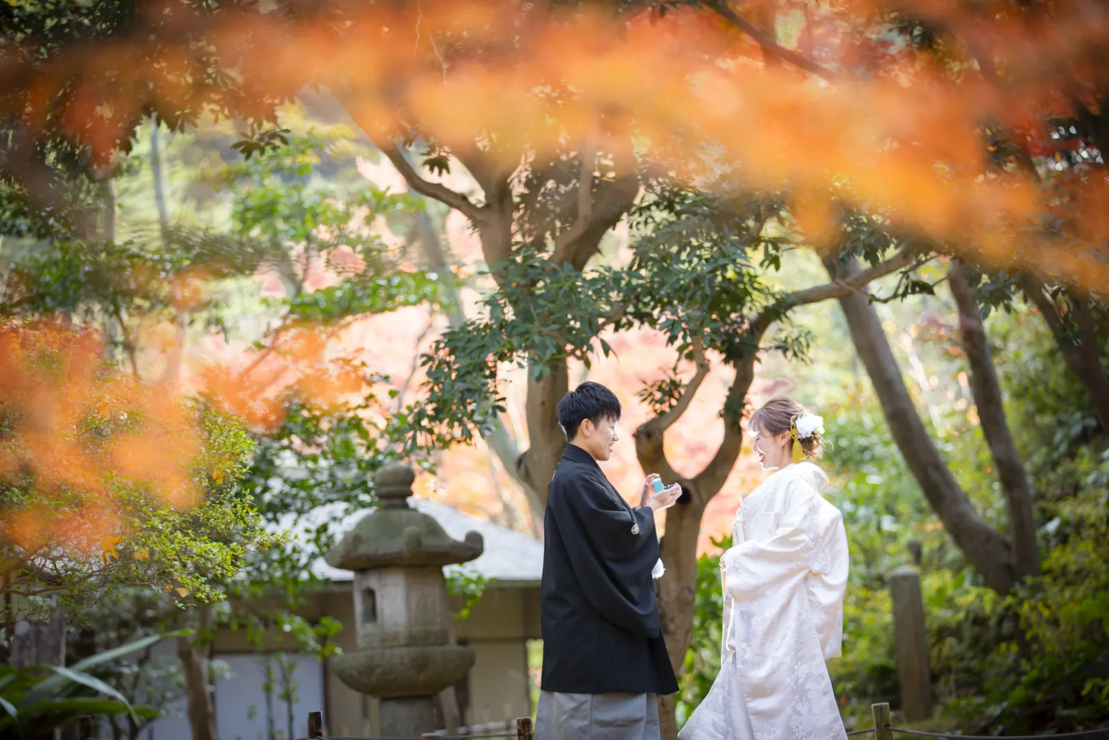 三溪園の色づいたもみじを前ボケに、石灯籠のそばで向かい合う白無垢の花嫁と紋付袴の新郎