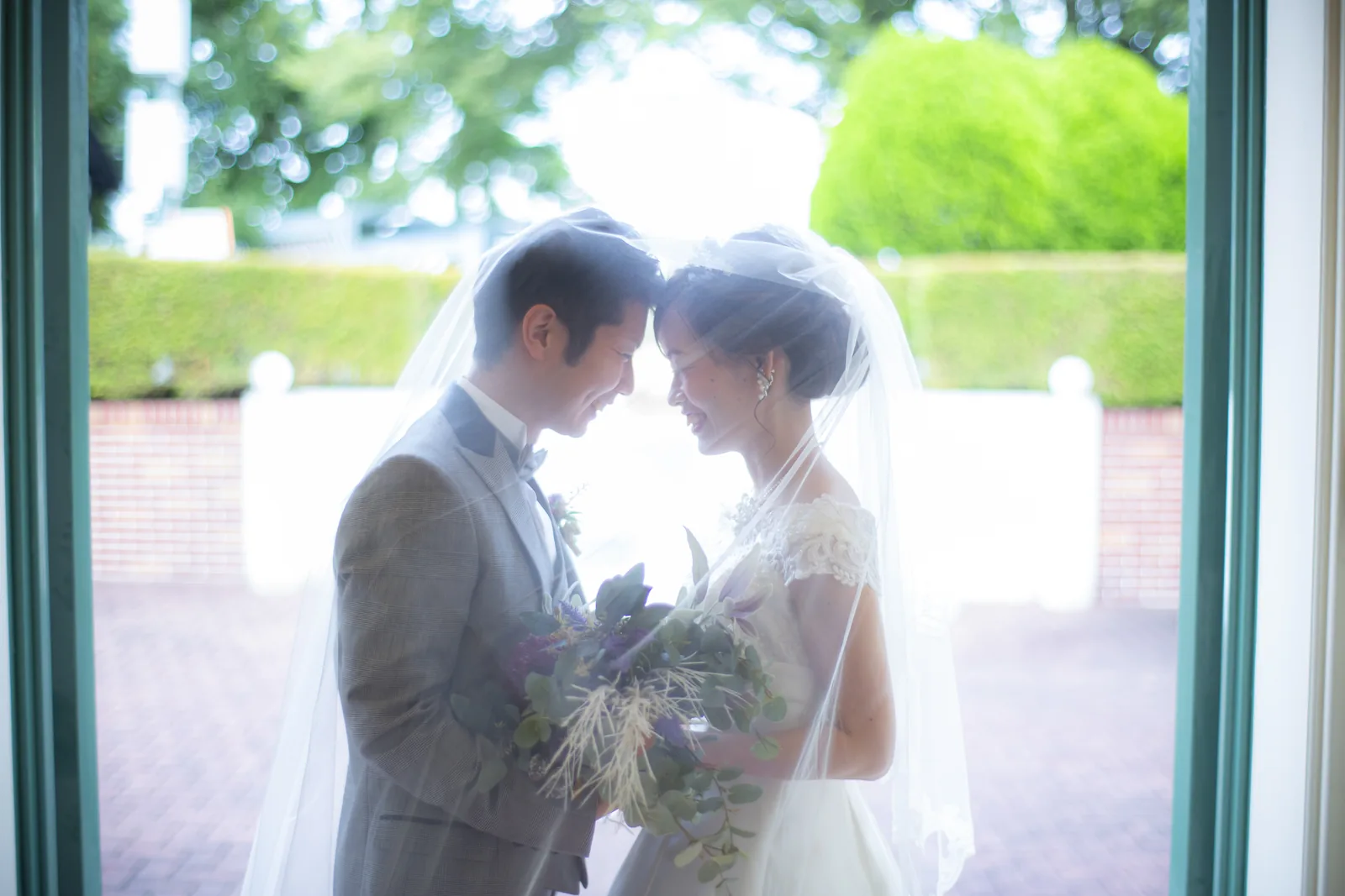 教会の緑を背景にベールの中で額を寄せる新郎新婦