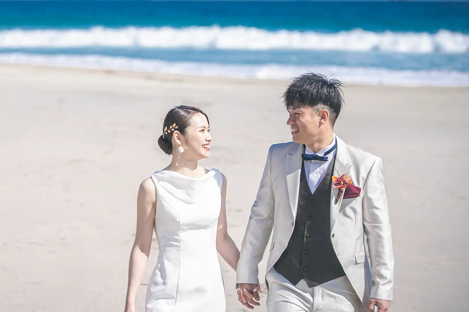 手をつないで笑顔で歩くウェディングドレスとタキシード姿の新郎新婦。青い海を背景にした明るく爽やかな洋装フォトウェディング。