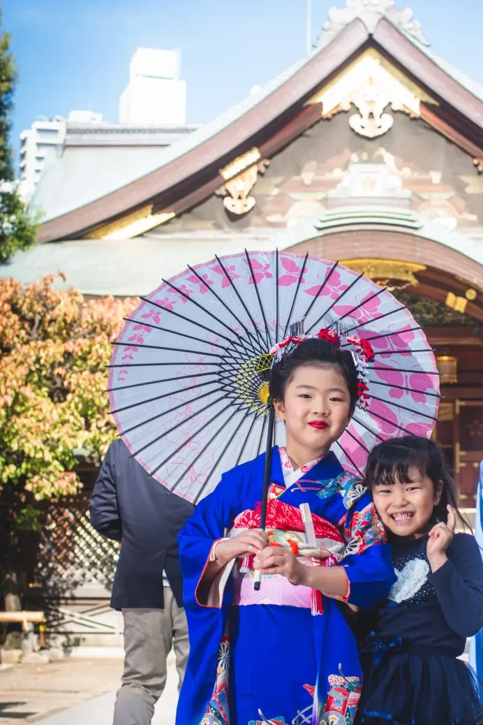 社殿の前でピンクの和傘を持つ7歳の女の子と洋服の妹、七五三の出張撮影（湯島天神・東京）